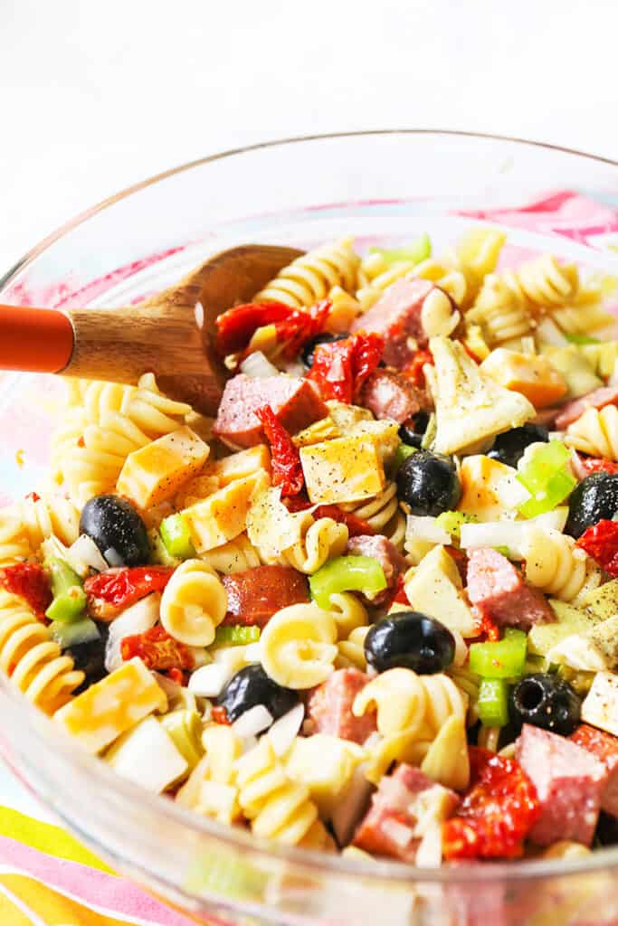 Pasta salad with a serving spoon scooping pasta, salami, cheese and more.