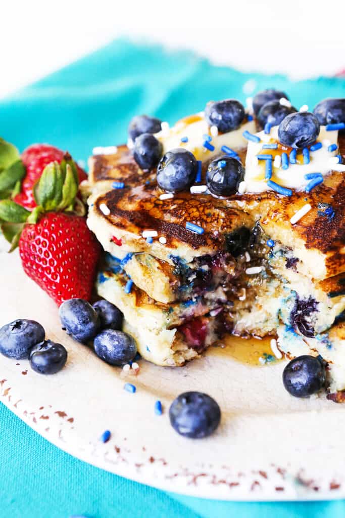 Pancakes without buttermilk in a stack with blueberries, strawberries and sprinkles on top with a bite removed. 