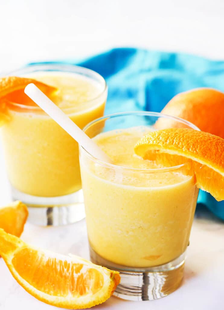 two glasses filled with orange julius beverage sitting next to oranges