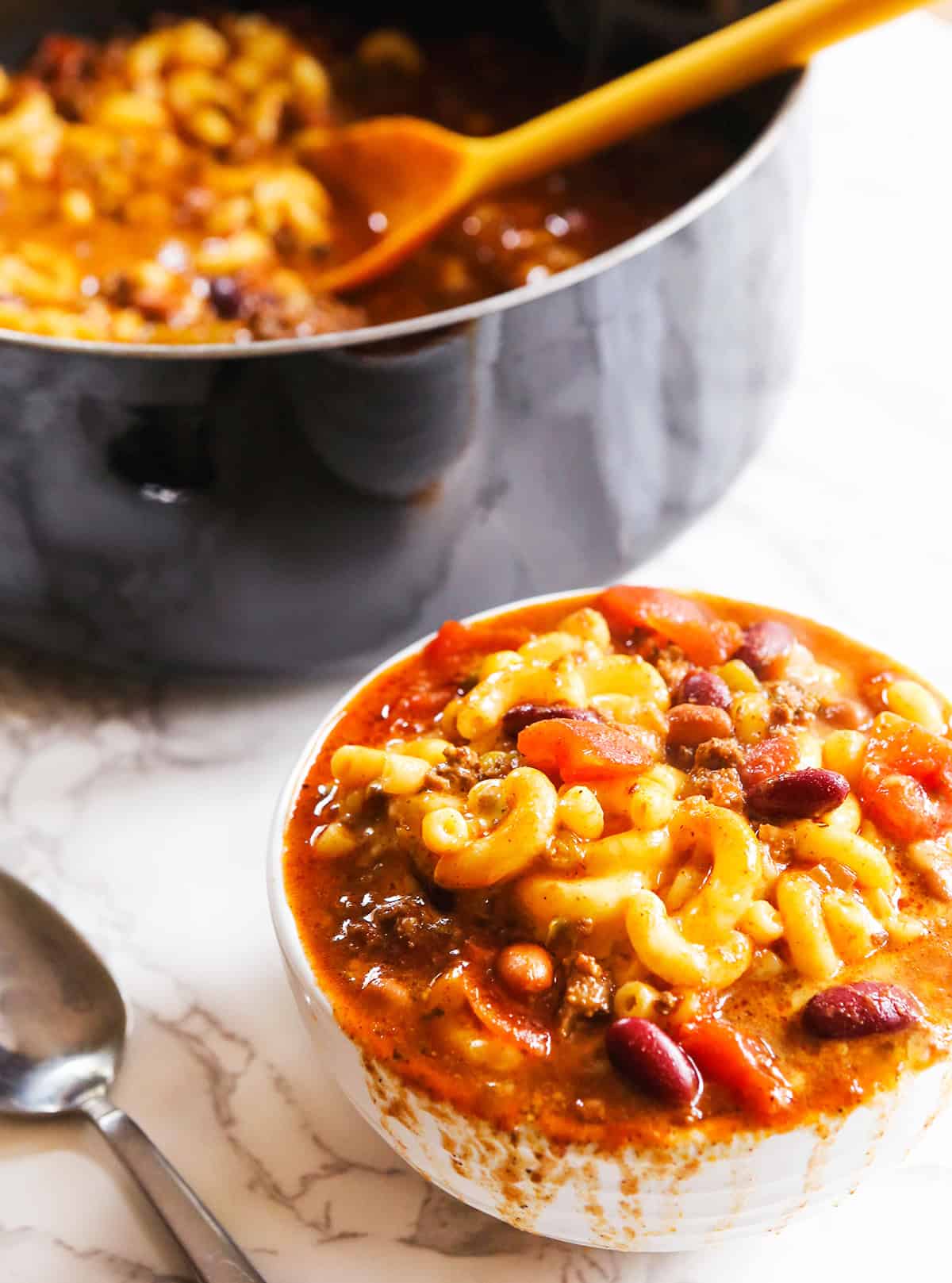 A heaping bowl of chili mac next to a large saucepan filled with it.