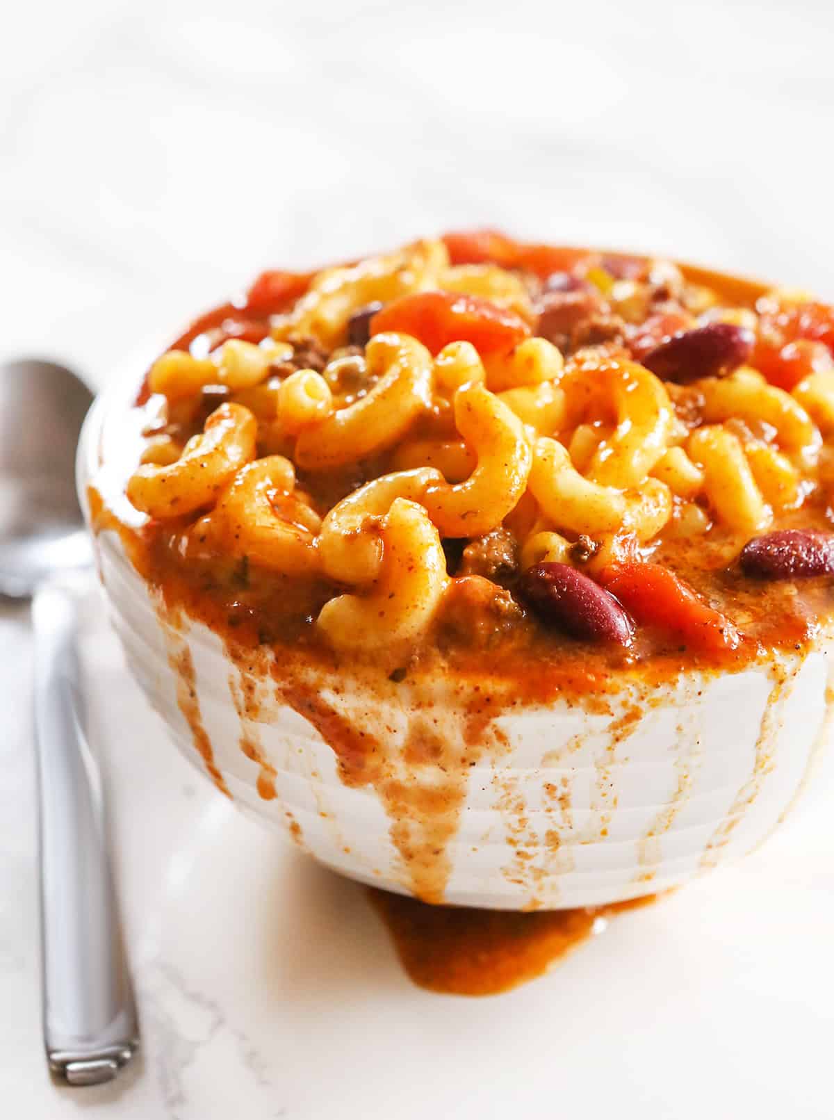 Heaping bowl of chili mac on a white counter, next to a spoon.