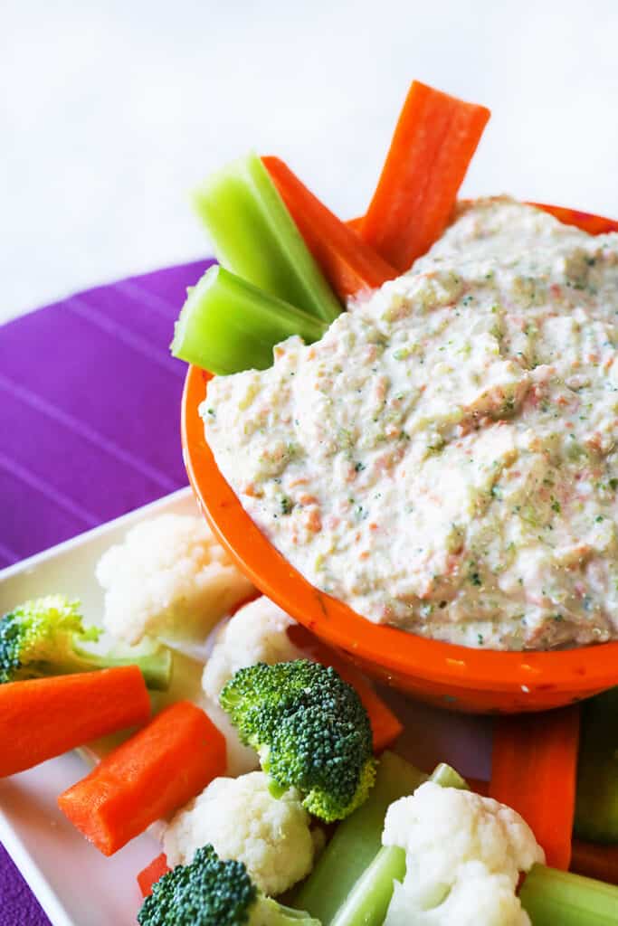 Creamy vegetable dip filled into a bowl and carrots and celery sticks tucked into it sitting on a platter of veggies. 
