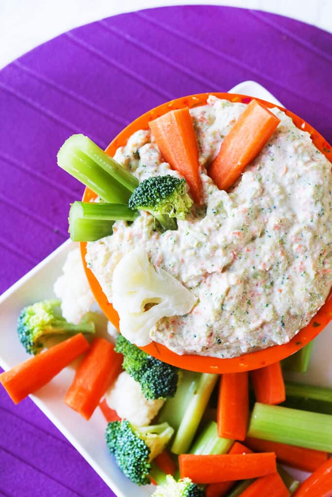 Bowl of creamy vegetable dip with carrots, celery and broccoli on top and around the bowl on a platter. 