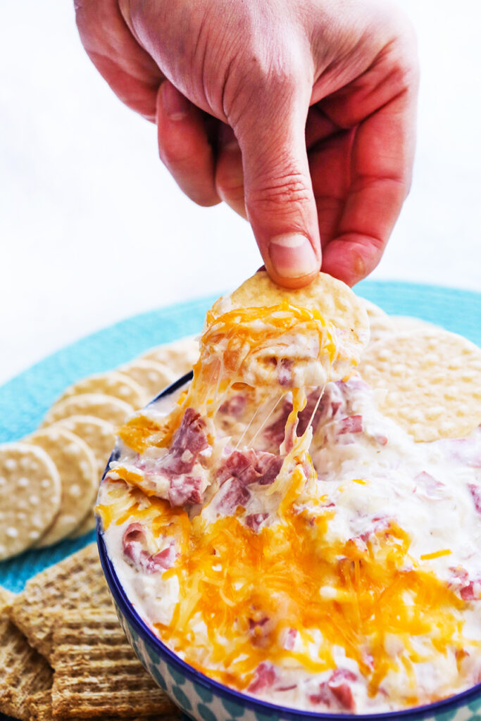 Hand pulling a cracker out of a serving bowl filled with cheesy reuben dip.