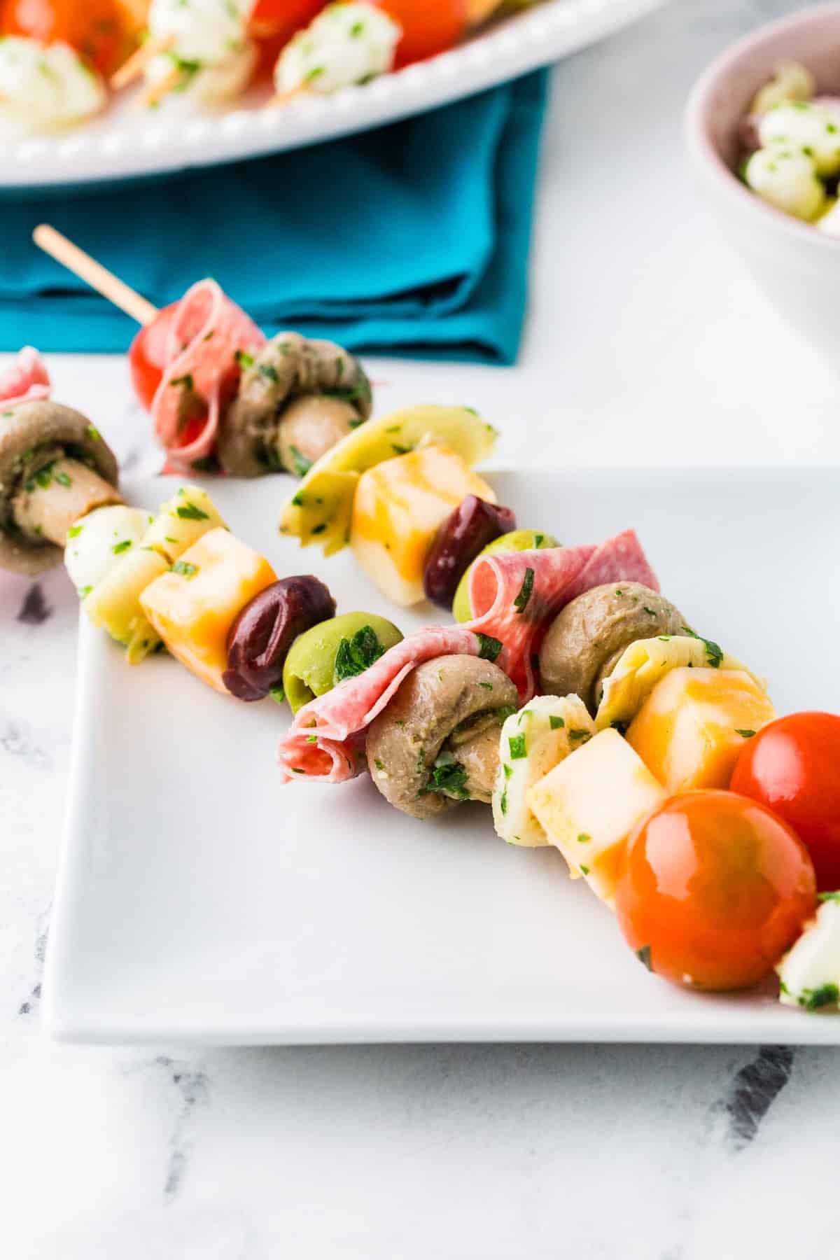 Two antipasto skewers on a small plate next to the serving plate with the rest of them on it.