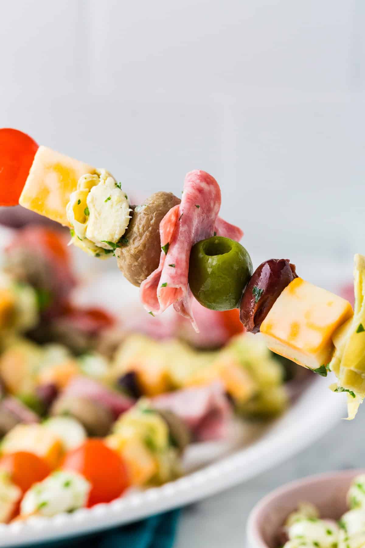 Loaded antipasto skewer being held up over a serving plate filled with more of them.