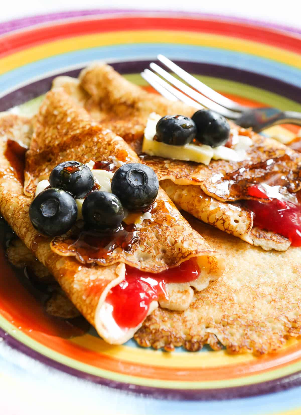 Two rolled up Swedish pancakes on a rainbow plate, topped with butter and blueberries.