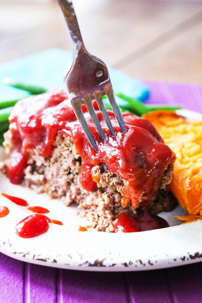 A fork sticking out of a slice of meatloaf with sauce dripping over it next to a halved sweet potato. 