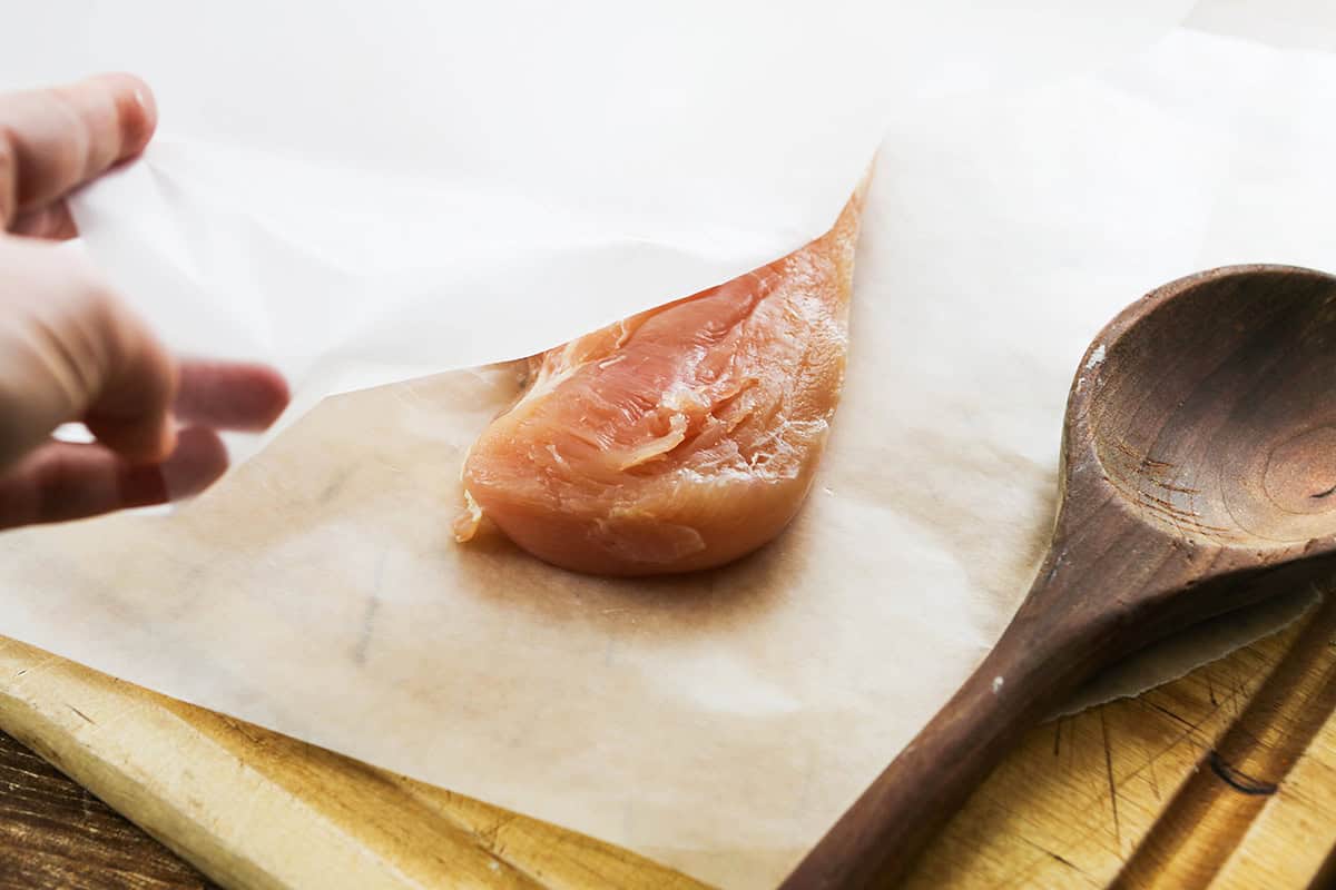 Hand revealing a pounded chicken breast between two pieces of wax paper.