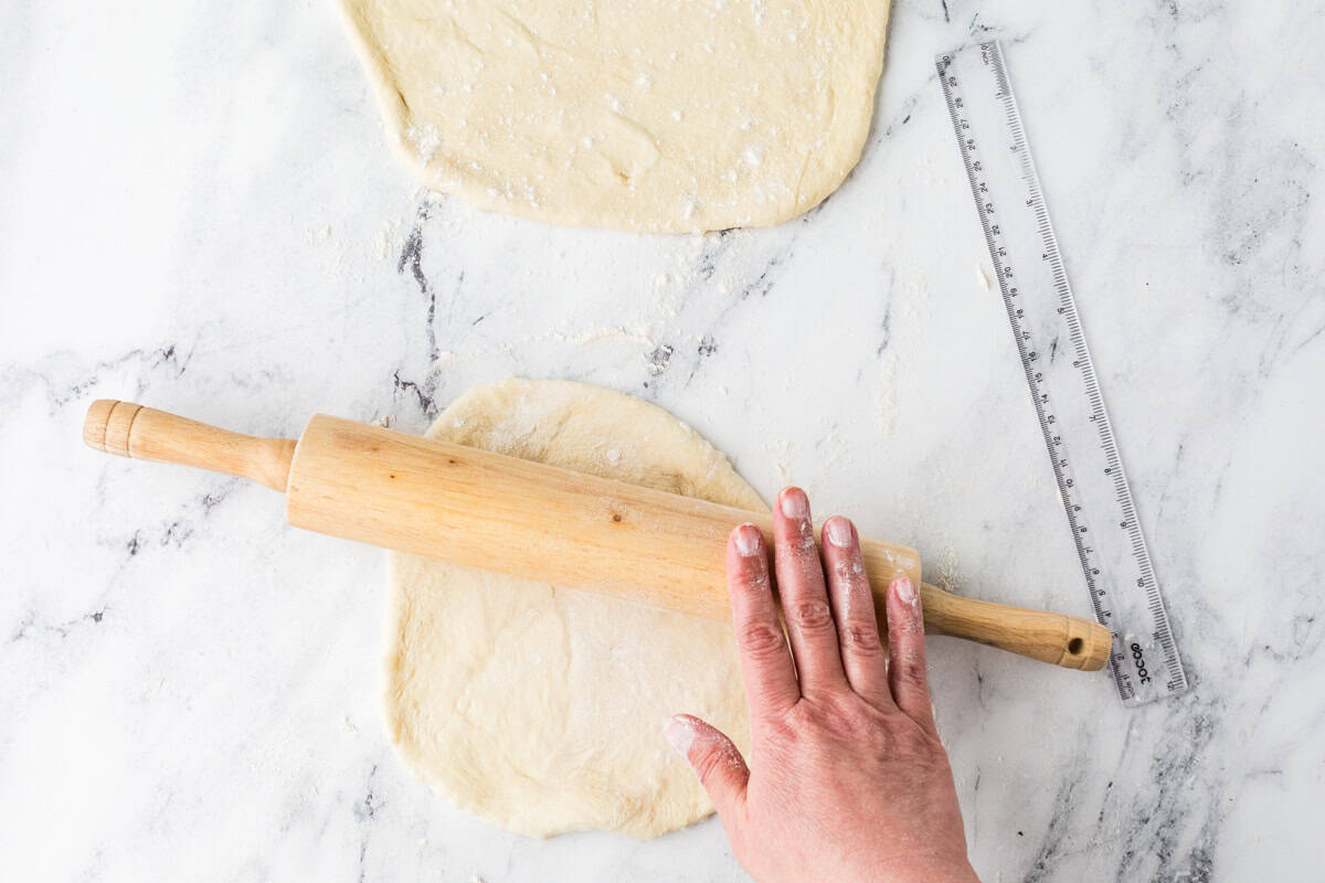 Hand rolling a rolling pin over a dough circle, next to another larger dough circle.