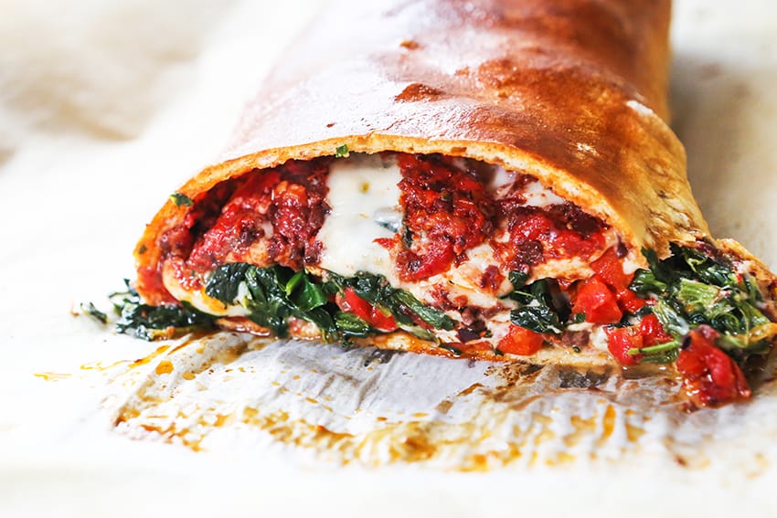 A veggie stromboli nicely browned and cut into so one end is oozing with cheese and veggies onto the cutting board. 