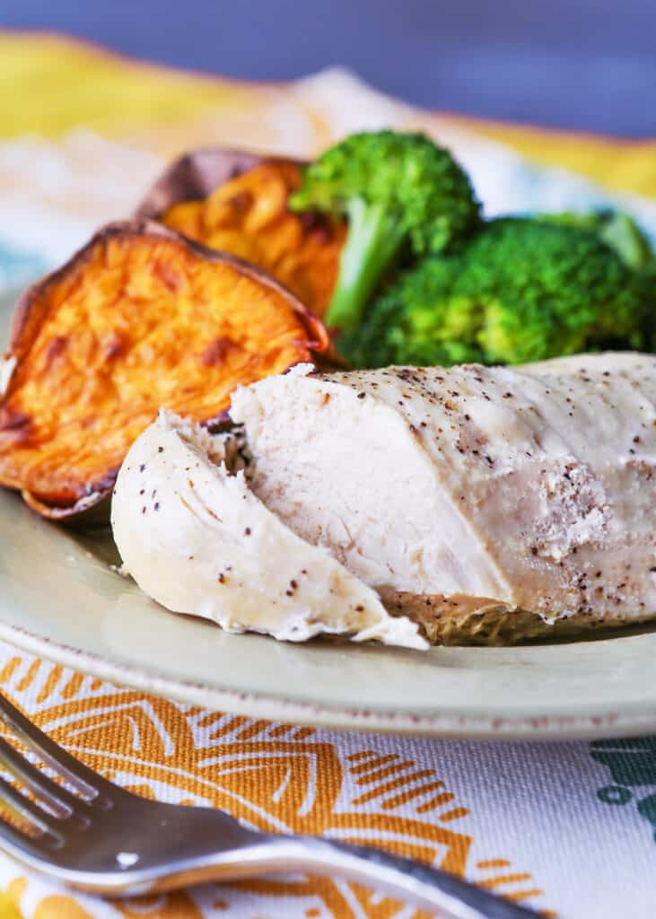 Plate with a grilled piece of chicken breast with a slice cut into it next to some broccoli and sweet potatoes. 