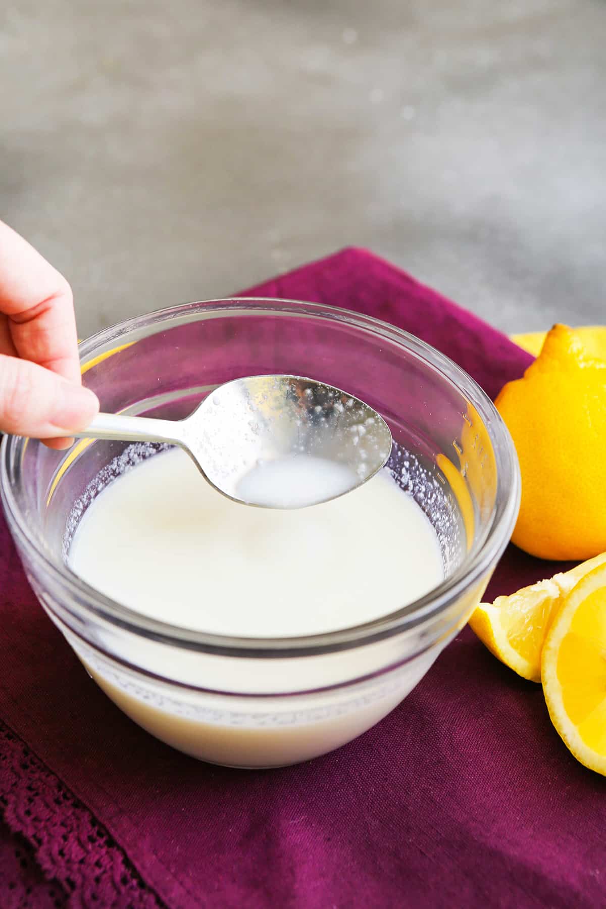 A spoon drizzling slightly chunky buttermilk into a bowl, sitting next to a lemon.