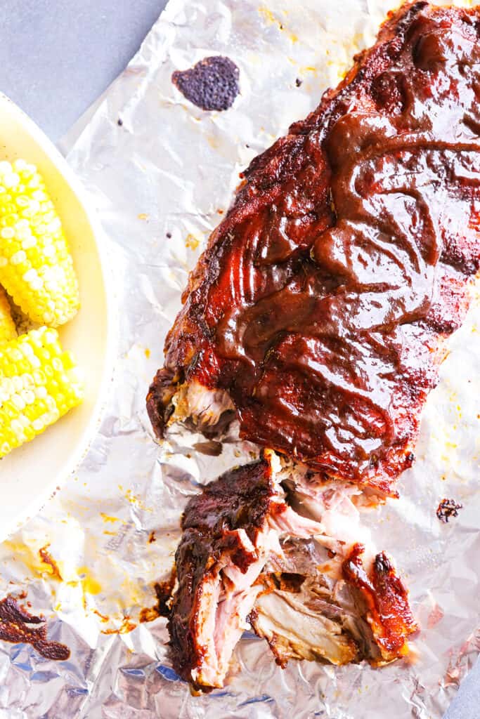 Rack of ribs on some tinfoil with a few slices removed sitting next to a plate of corn on the cob. 