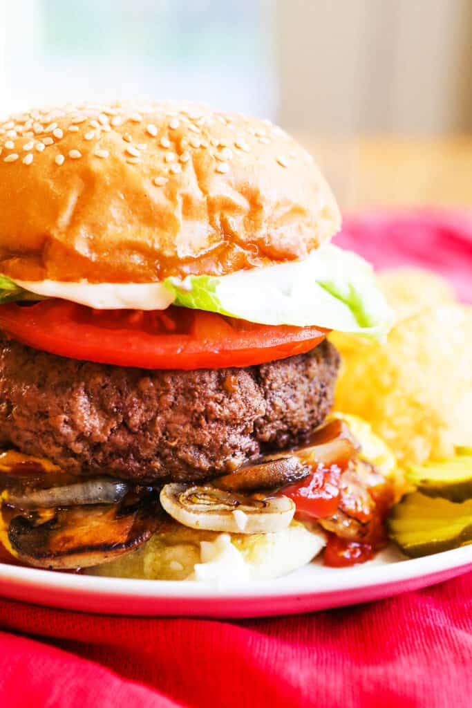 A five guys burger topped with lettuce and tomato on a plate with grilled onions, mushrooms and pickles next to some chips. 