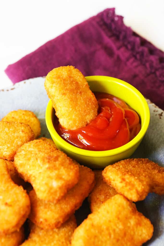 Air fried chicken nuggets on a plate with a bowl of ketchup that has a nugget sitting in it. 