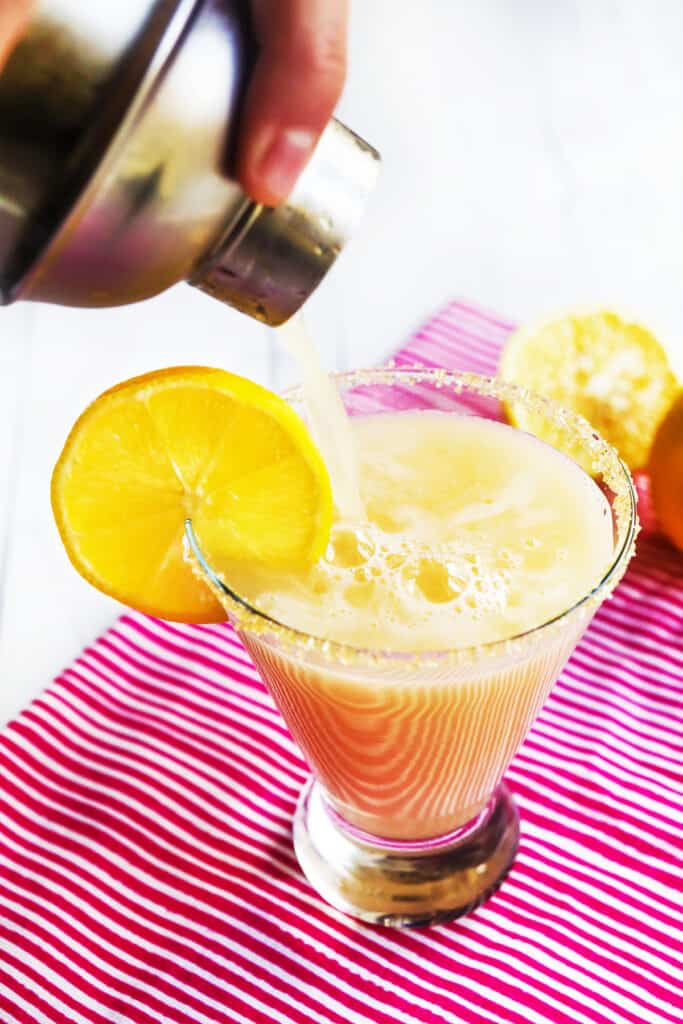 Hand holding a cocktail shaker  pouring a lemon drop icocktail nto a highball with a lemon garnish on the side. 