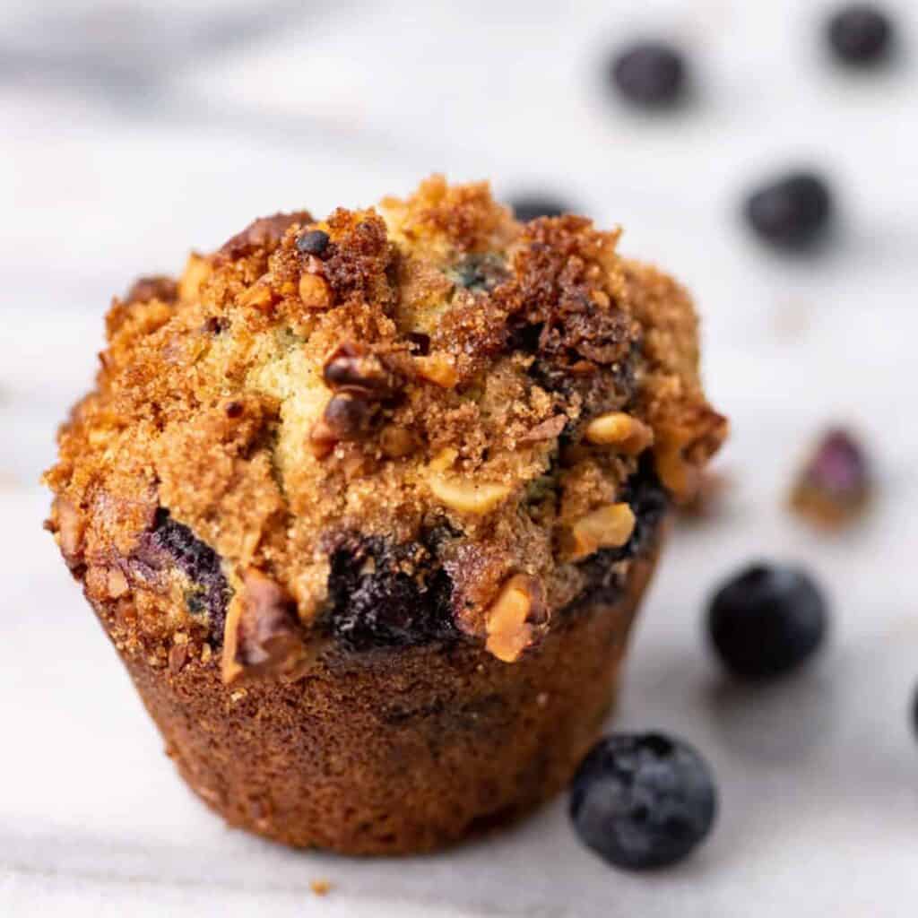 single blueberry muffin sitting on counter next to fresh blueberries