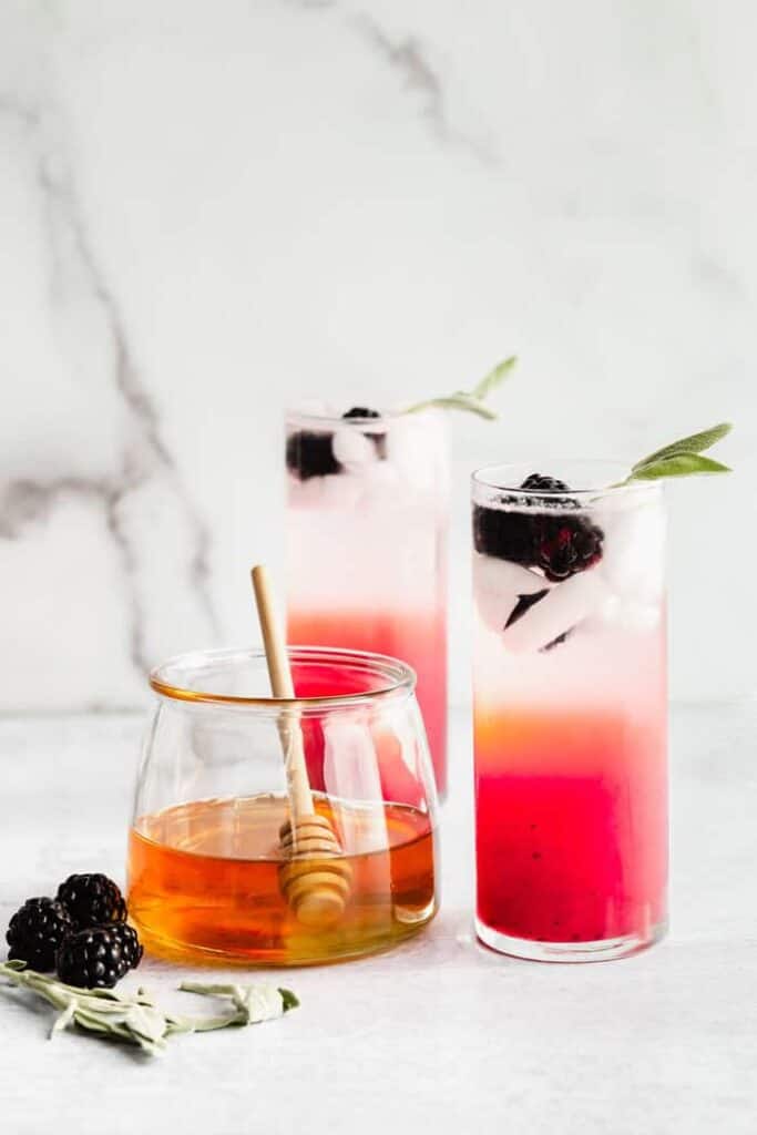 Honey, blackberry, sage refresher mocktail with a sage garnish sitting next to a pot of honey.