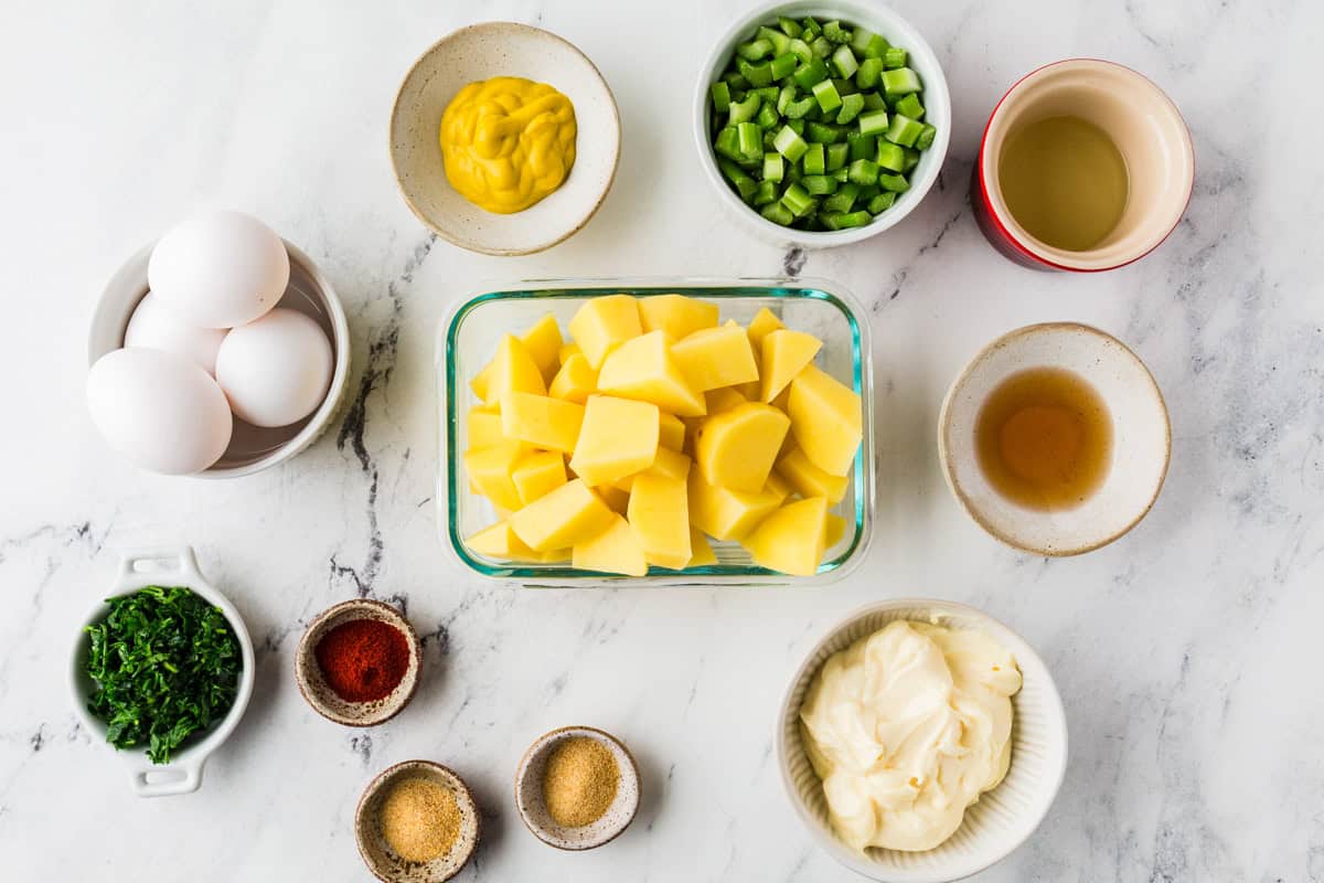 Ingredients for potato salad in individual bowls lined up on a white counter.