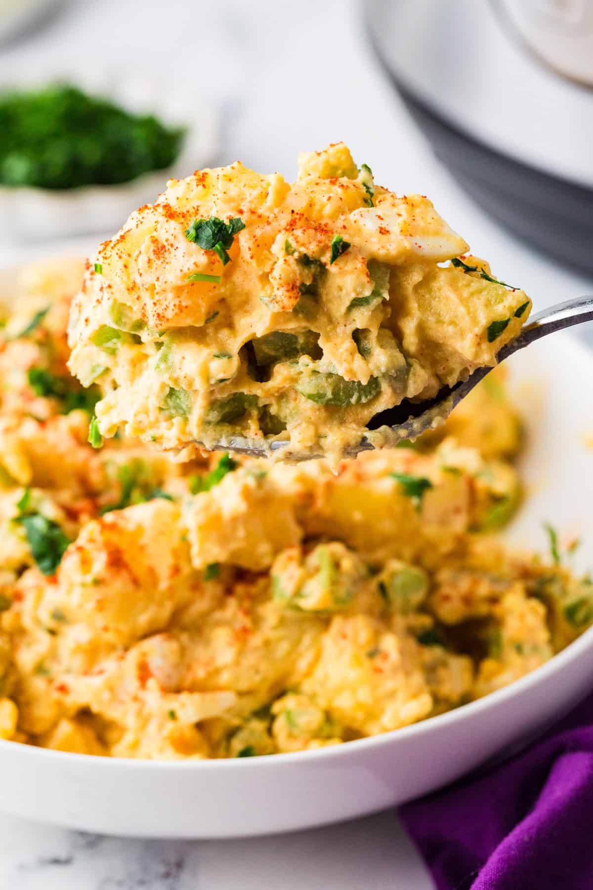 Serving spoon holding up a spoonful of potato salad, hovering over a plate filled with the mixture.