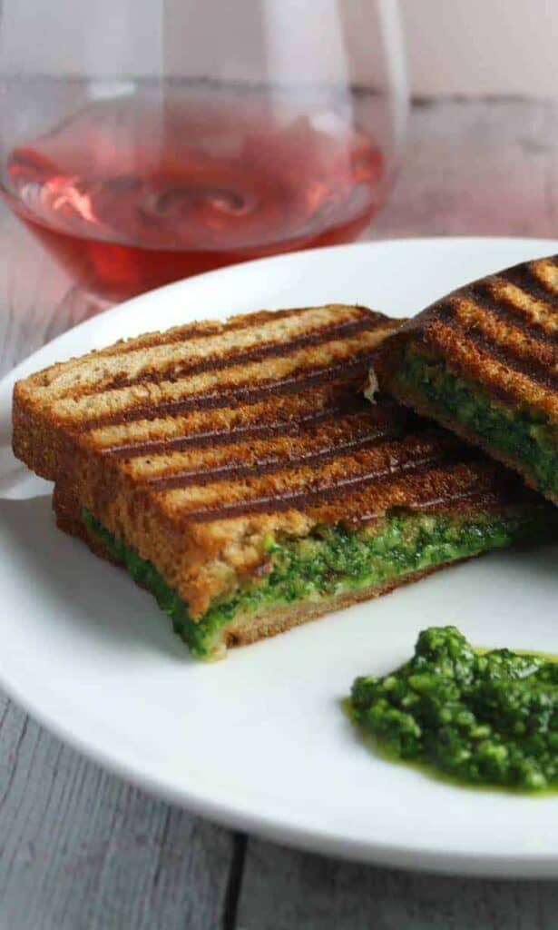 a kale pesto grilled cheese sandwich with grill marks on top cut in half sitting on a plate next to a glass of wine