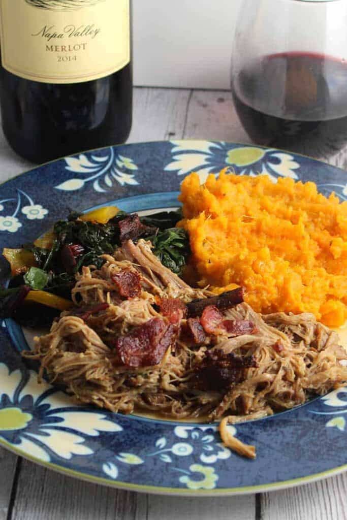 pulled pork with bacon shredded on  a plate with a mashed sweet potato on the side and a glass of wine off to the side