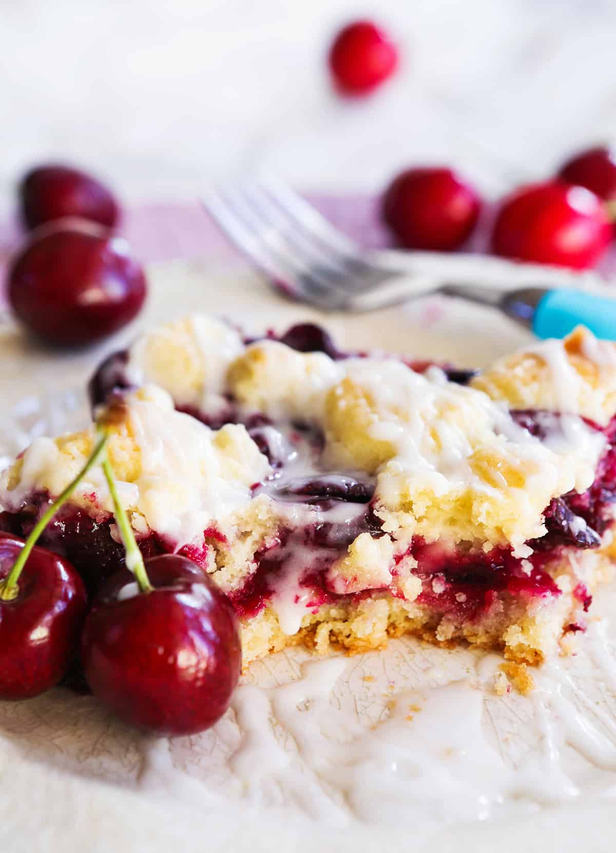 A piece of cherry bars with a bite taken out on a plate surrounded by cherries.