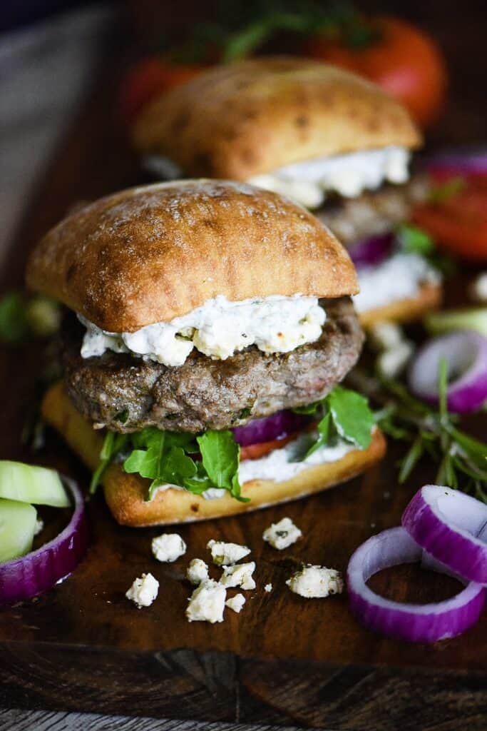 Feta crumbles and red onion sitting in front of a lamb burger on a ciabatta bun with Tzatziki sauce.