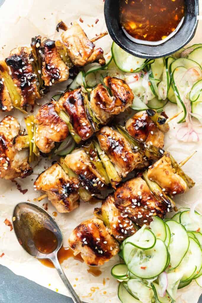 Chicken teriyaki skewers on a platter with a spoon with leftover sauce that was drizzled over the chicken and pickled cucumbers on the side. 