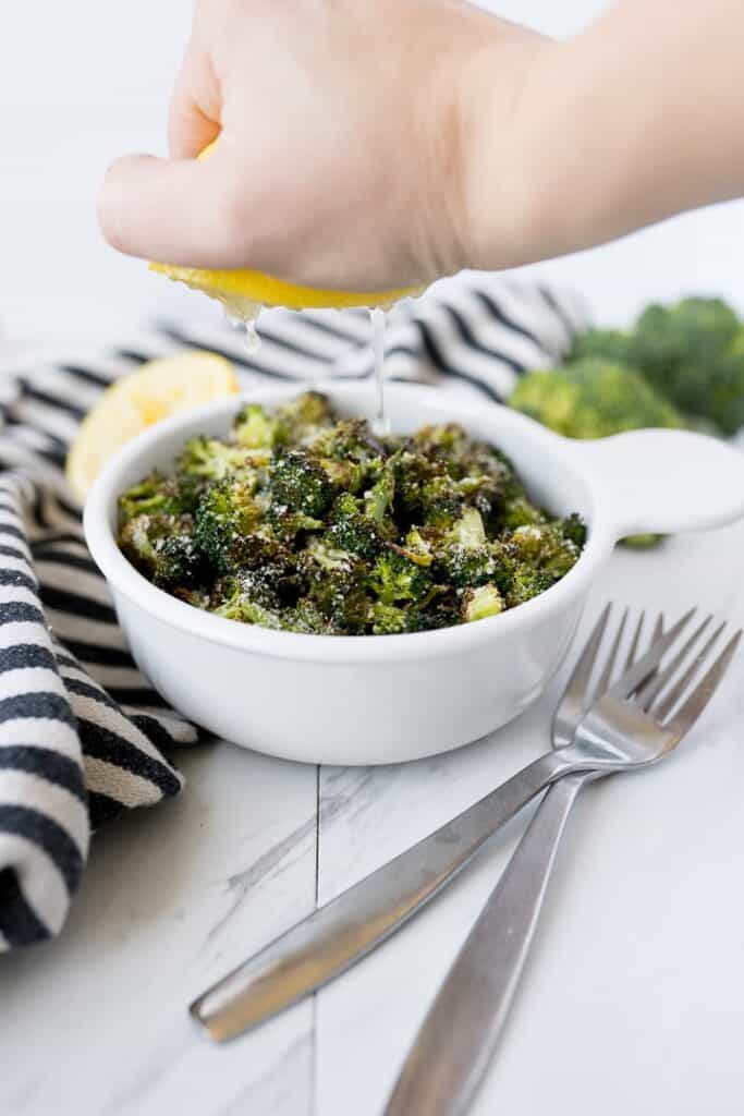 Roasted lemon parmesan broccoli in a bowl with a hand squeezing fresh lemon juice over the serving bowl.
