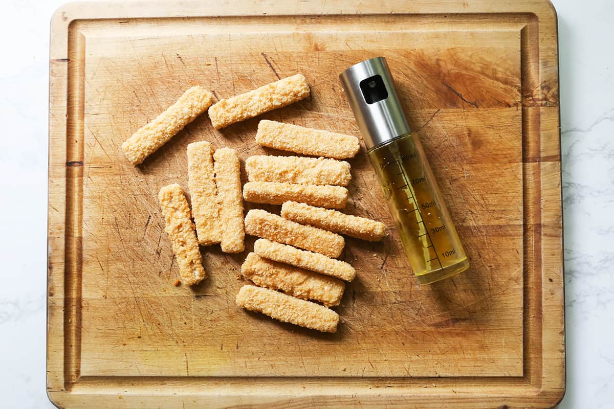 Frozen fish sticks on a cutting board with a spritzer filled with olive oil.