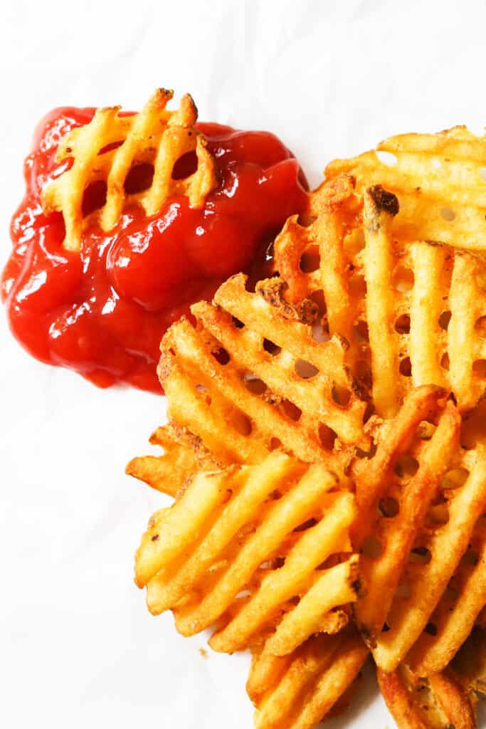 Waffle fries in a pile, next to ketchup on parchment.