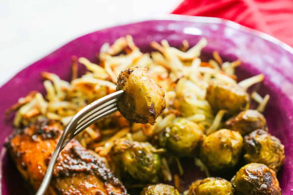 Fork holding a single Brussels sprout over a plate of salmon, shredded hash browns and Brussels sprouts. 