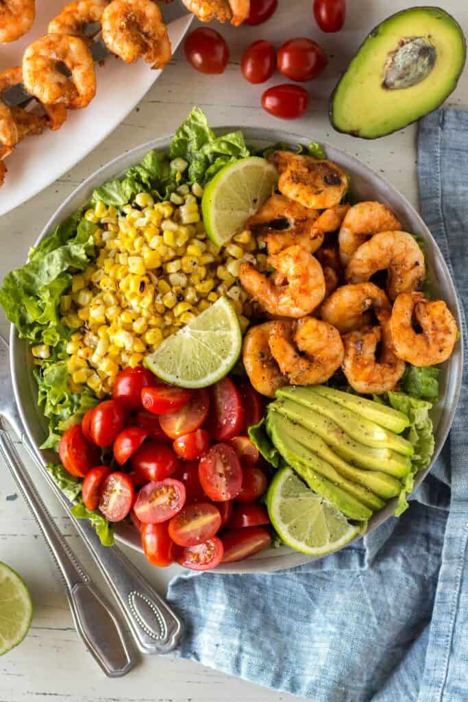 grilled shrimp, corn, avocado and tomatoes sittong on a bed of lettuce with serving utensils next to it and lime wedges on top. 