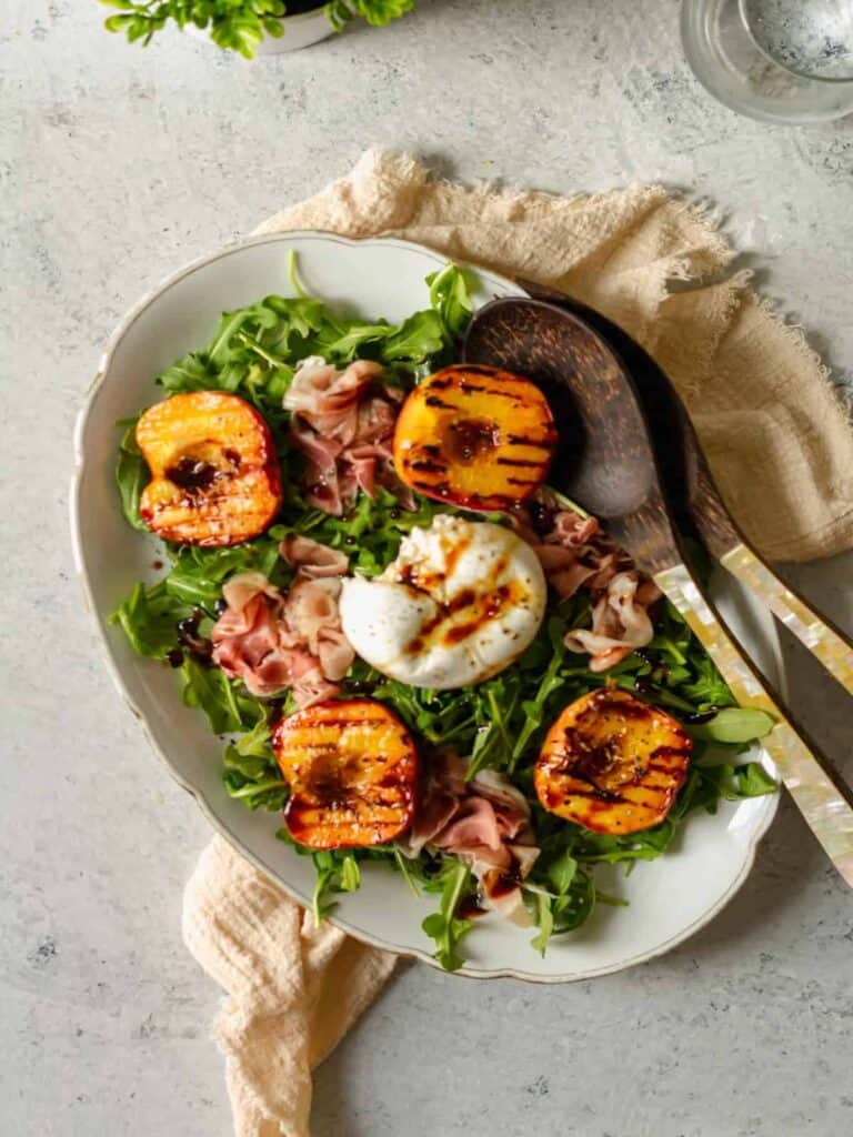 Serving platter of prosciutto, burrata and grilled peach salad with serving spoons inside. 