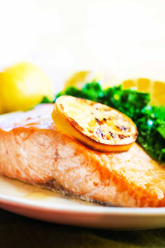 Slice of salmon on a plate with greens and a lemon garnish on top. 