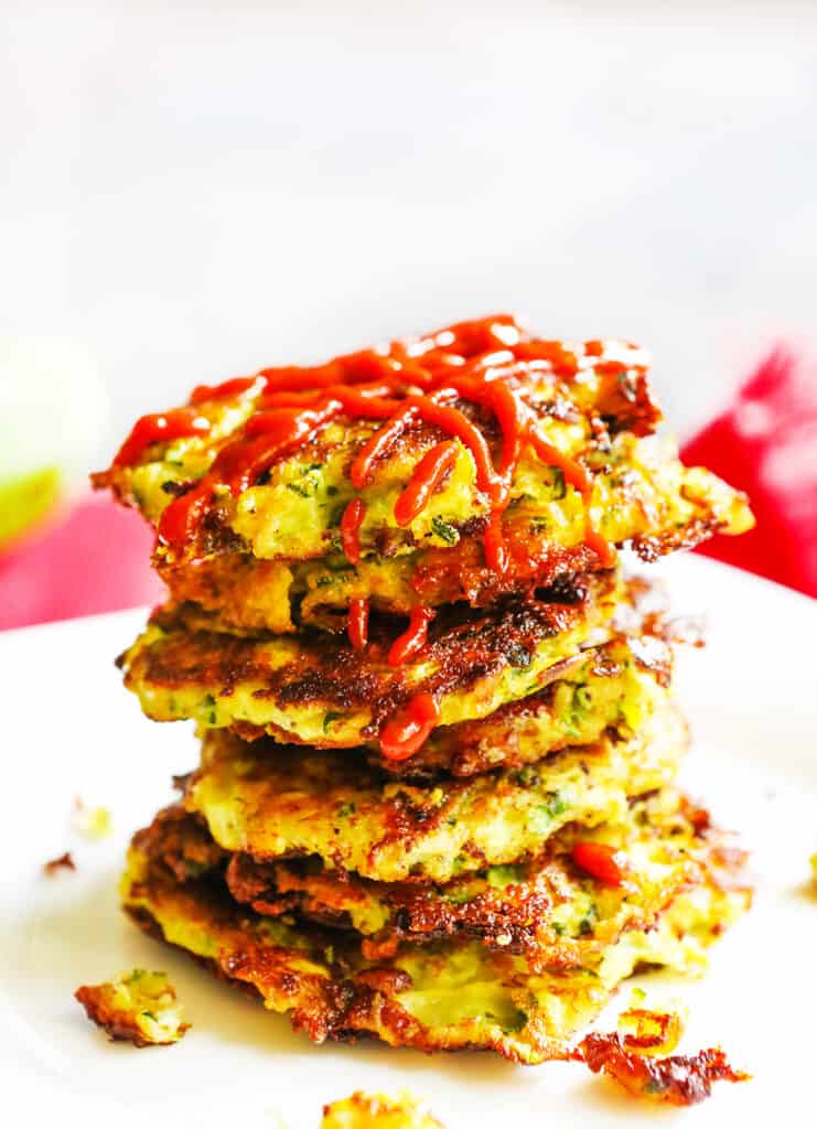 Stack of six zucchini fritters on a plate with ketchup drizzled over the top. 