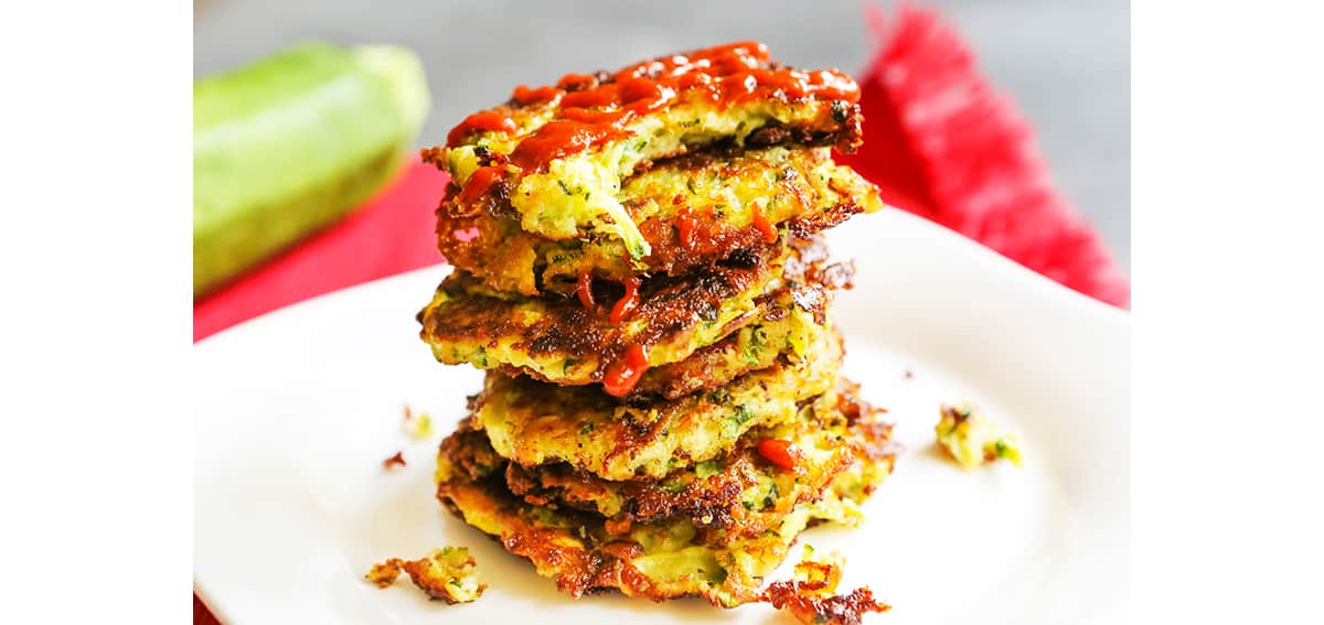 Stack of zucchini fritters on top of one another and sriracha drizzled over the stack.