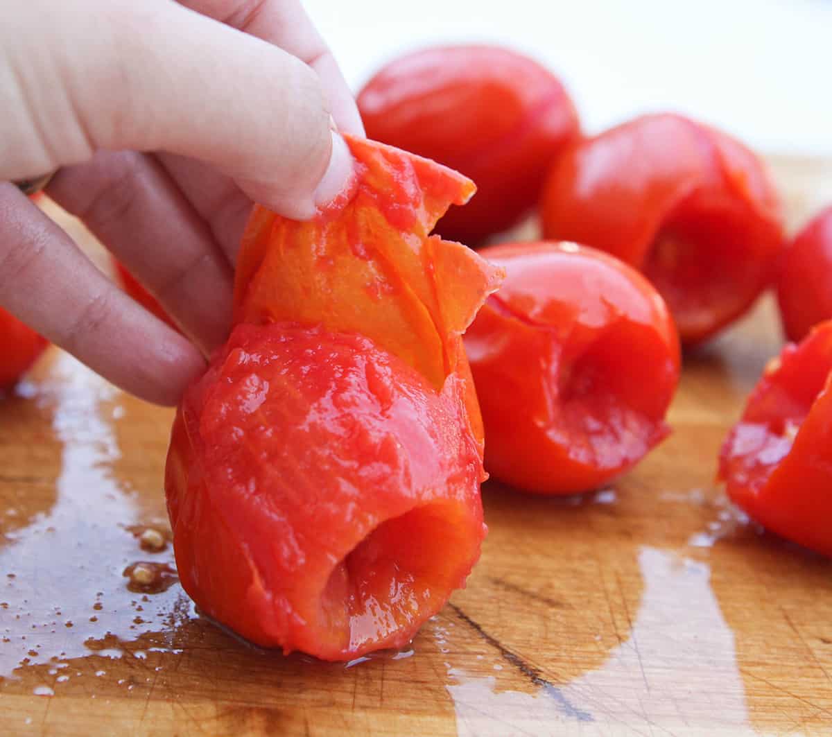 Fingers peeling skin off tomato on cutting board.