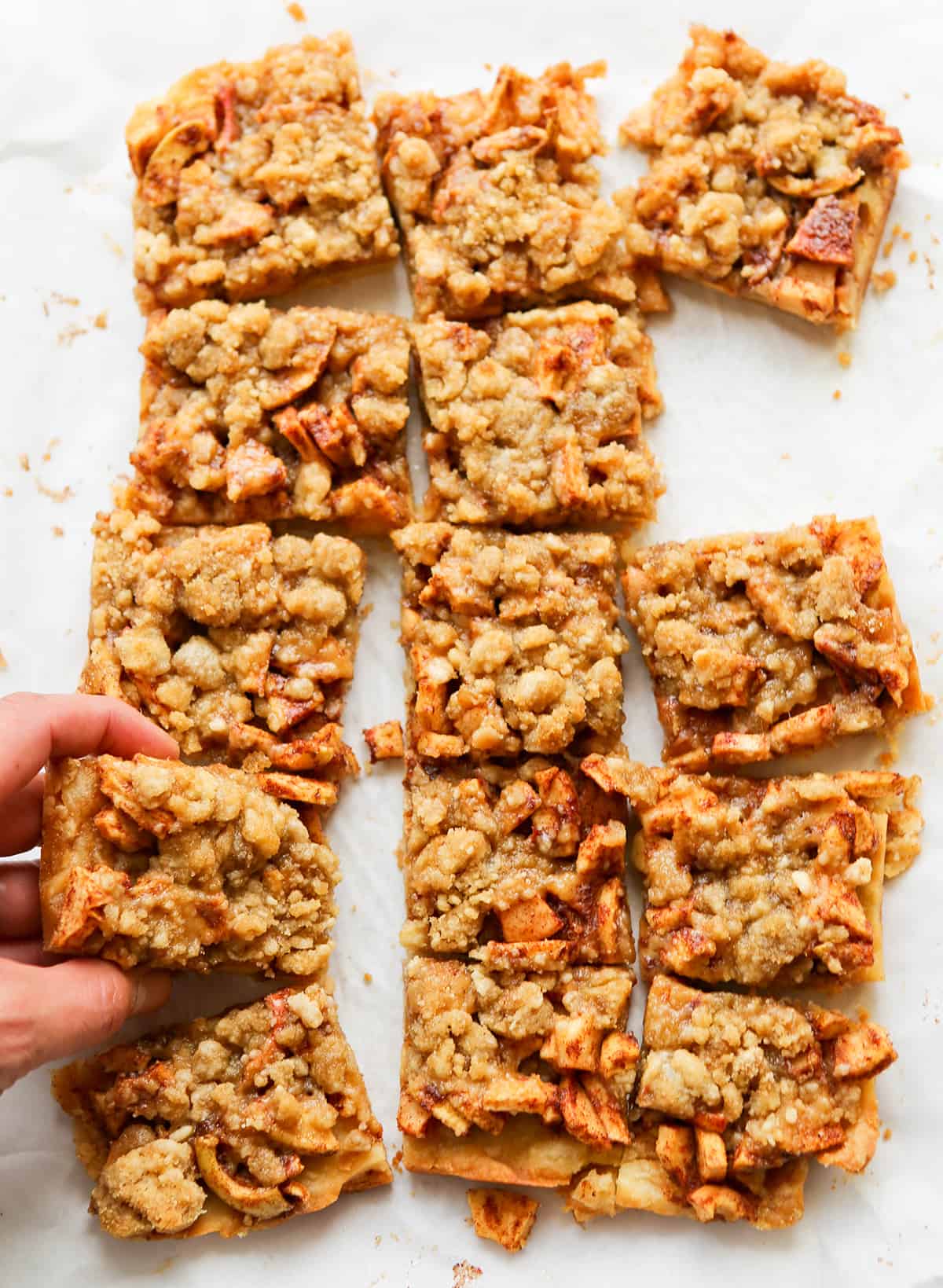 Apple pie bars lined up on parchment paper, with a hand taking one away.