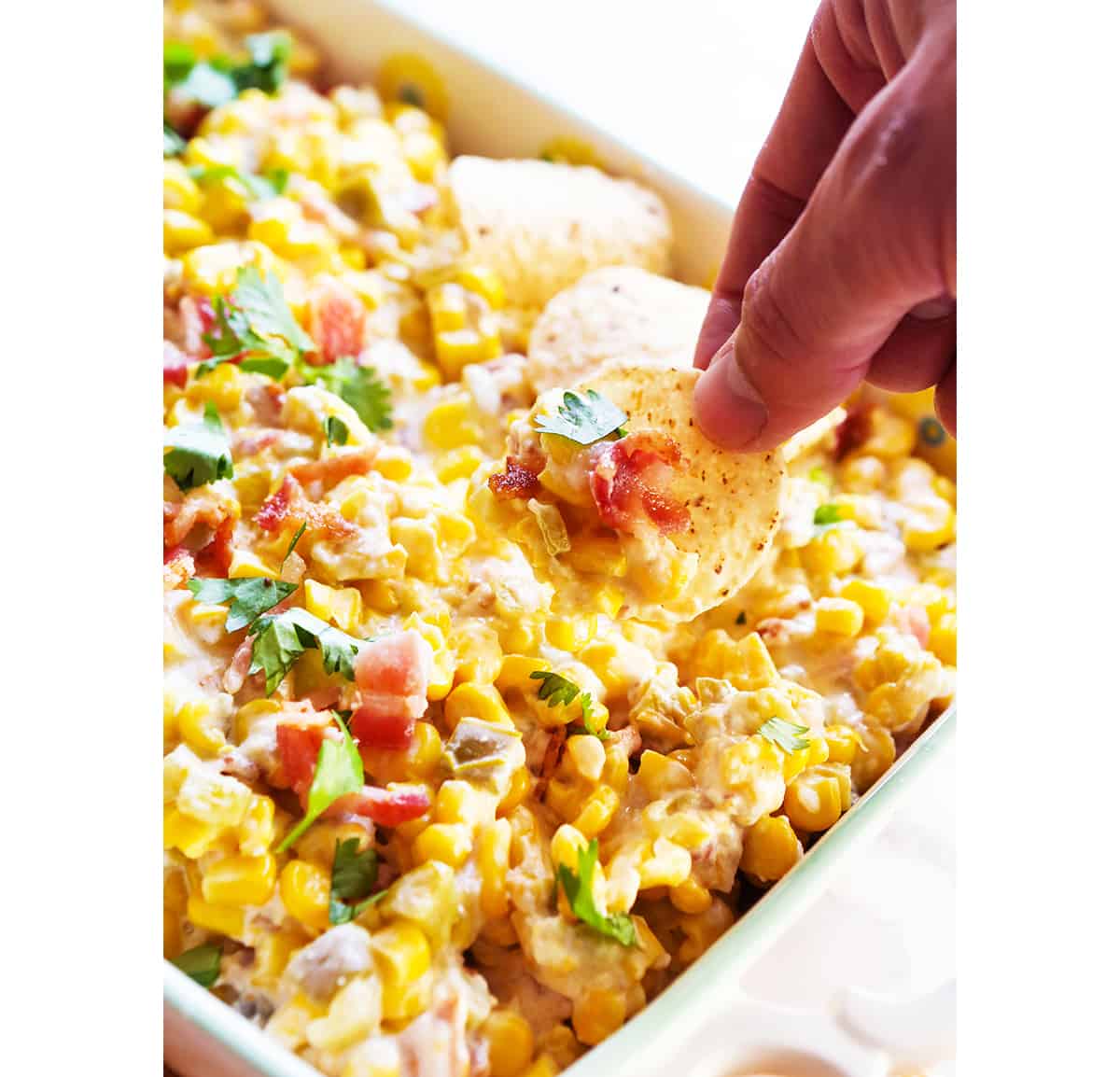 Hand with tortilla chip scooping corn dip out of baking dish.
