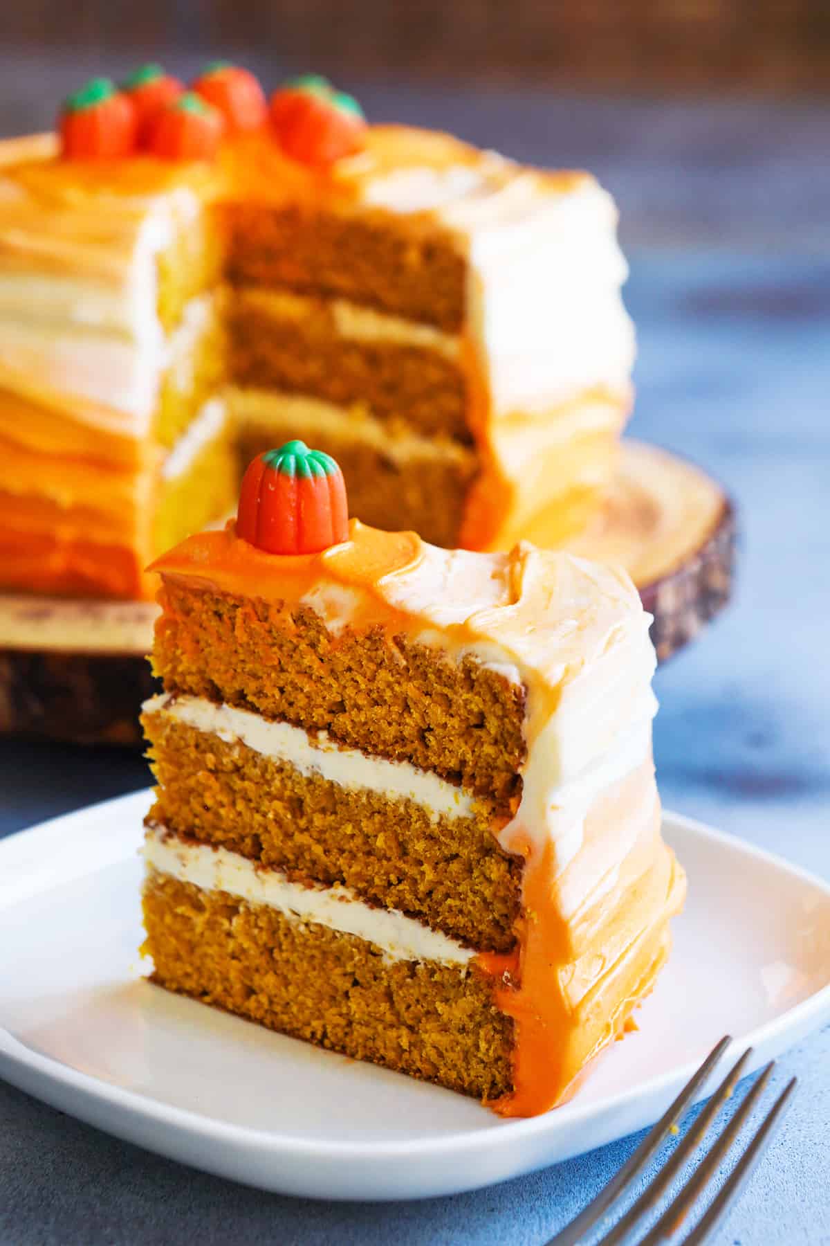 A slice of pumpkin spice layer cake on a plate with the full cake behind it off to the side. 
