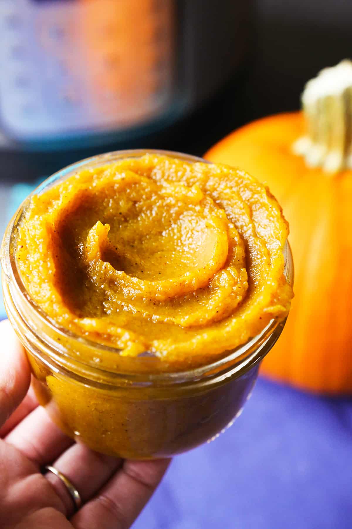 Hand holding a jar of homemade pumpkin puree, with a pumpkin and pressure cooker in the background.