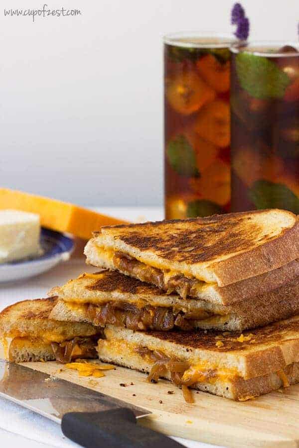 Apple cider vinegar caramelized onion grilled cheese sandwiches cut diagnolly and stacked on top of one another on a plate next to a couple of glasses of ice tea. 