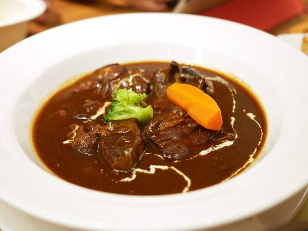 Beef tongue stew portioned into a soup bowl with a carrot and broccoli garnished on top. 