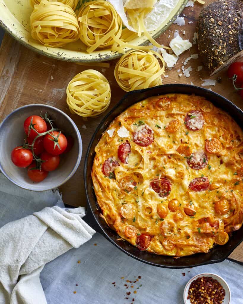 Cast iron skillet of leftover pasta frittata on a table with fresh tomatoes and uncooked pasta balls sit near it. 