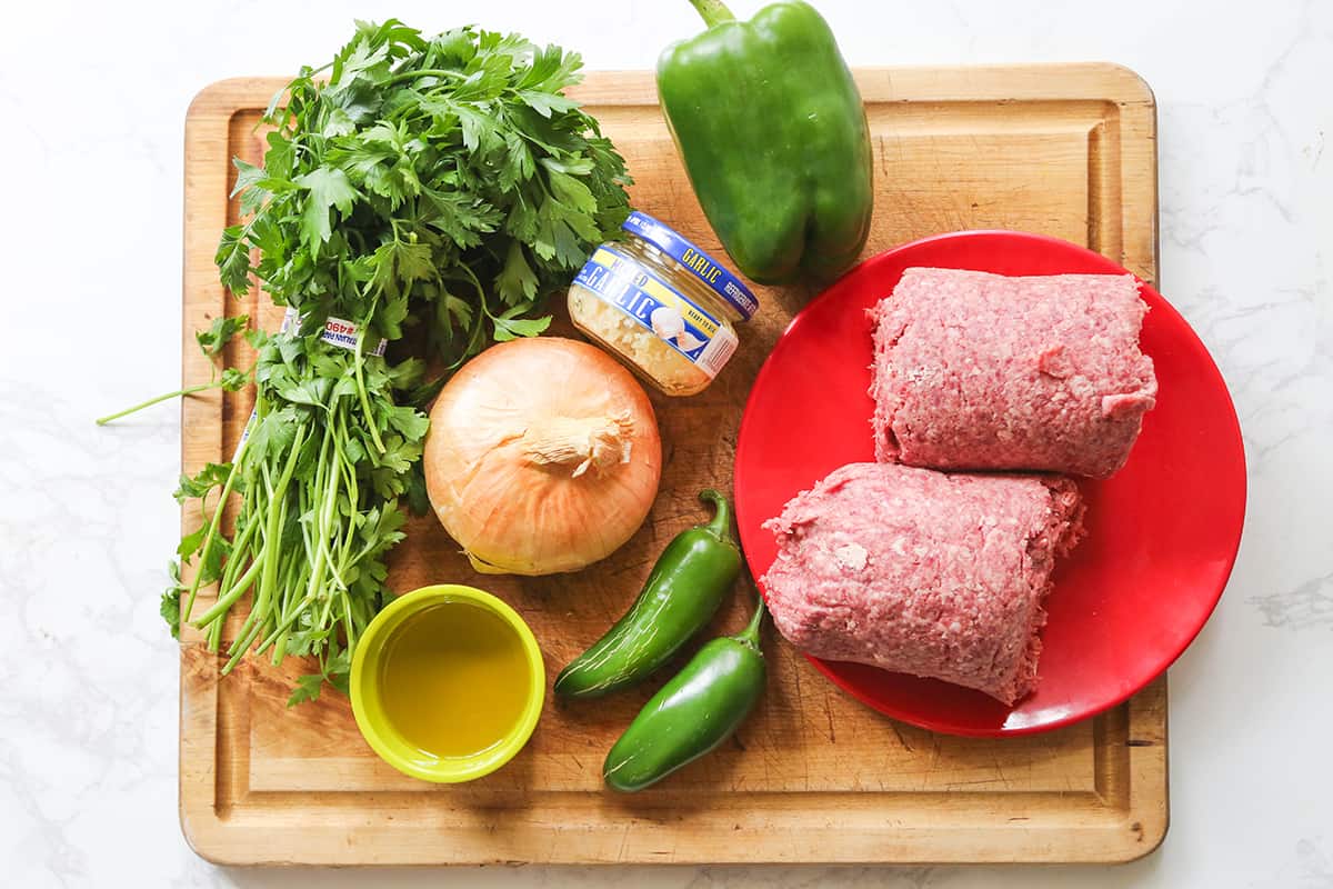 Ground beef on a cutting board with parsley, bell pepper and other ingredients.