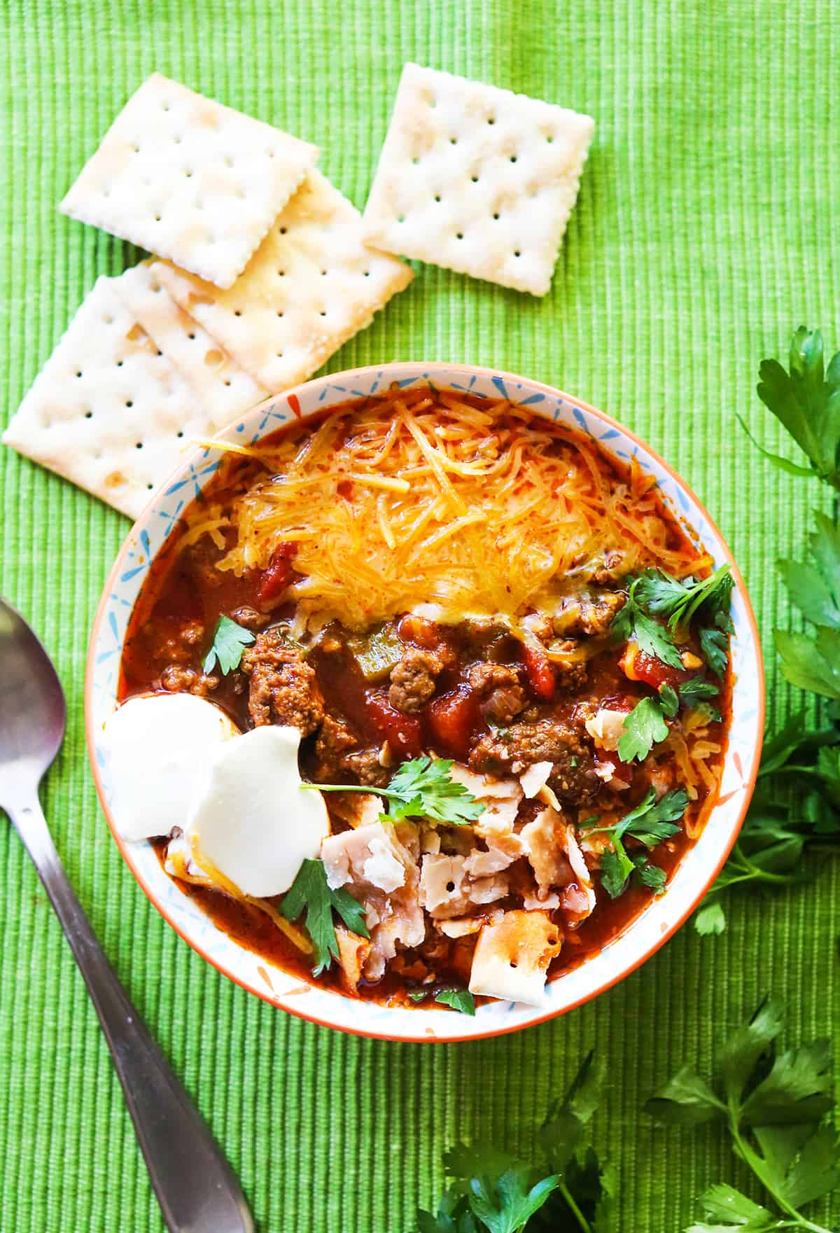 Looking down into a heaping bowl of chili without beans topped with sour cream, crackers and parsley.