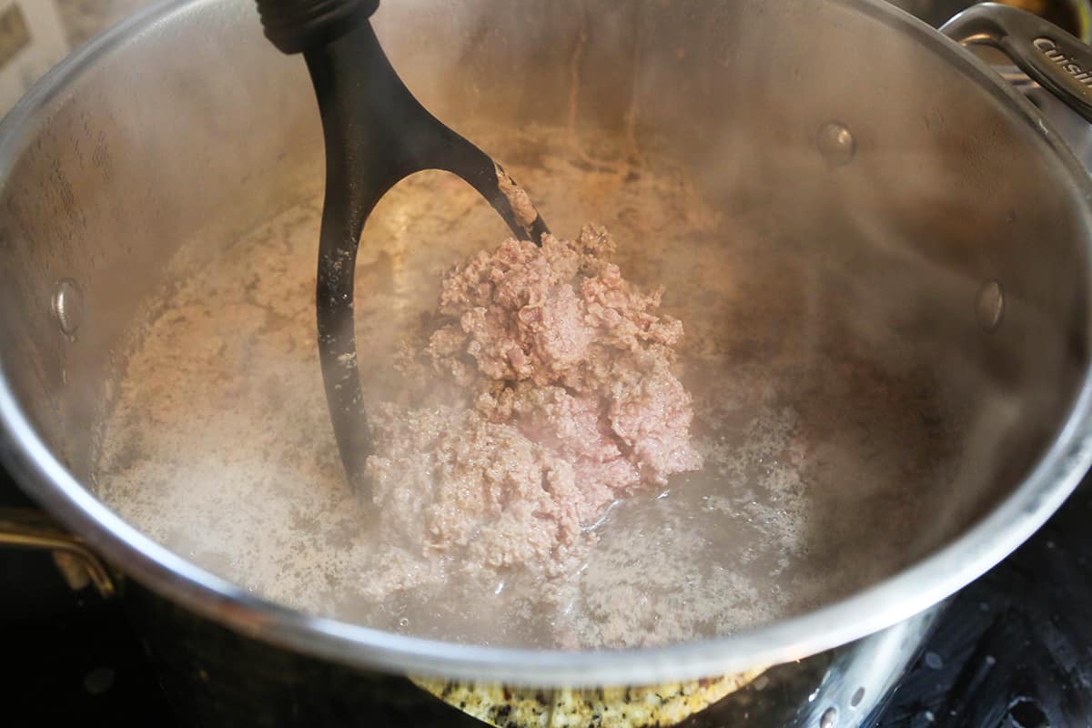 Potato masher mashing ground beef in boiling water inside a large saucepan.